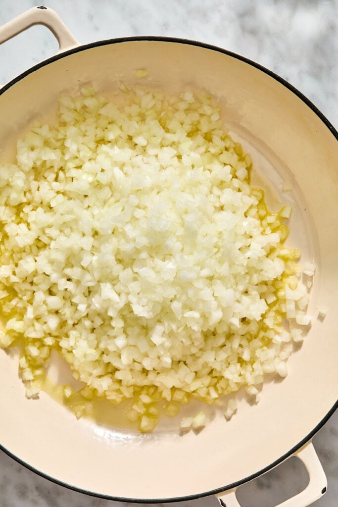 Finely diced onions cooking in oil in a light-colored enameled cast iron pan.