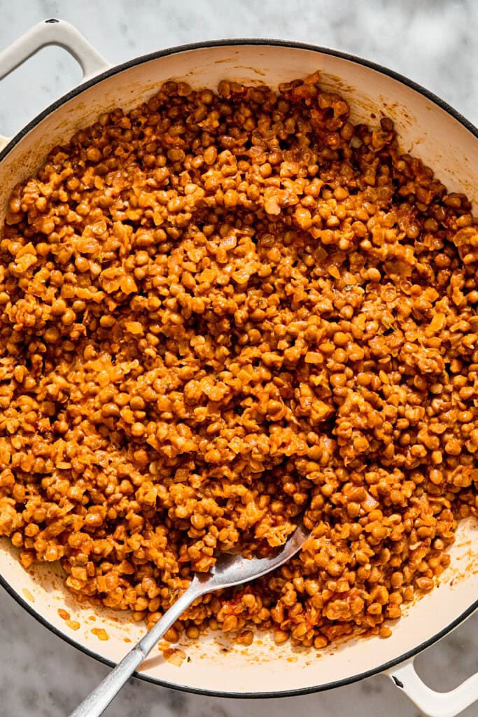 A white enameled cast iron skillet filled with cooked brown lentils in a tomato-based sauce, with a metal spoon resting inside.