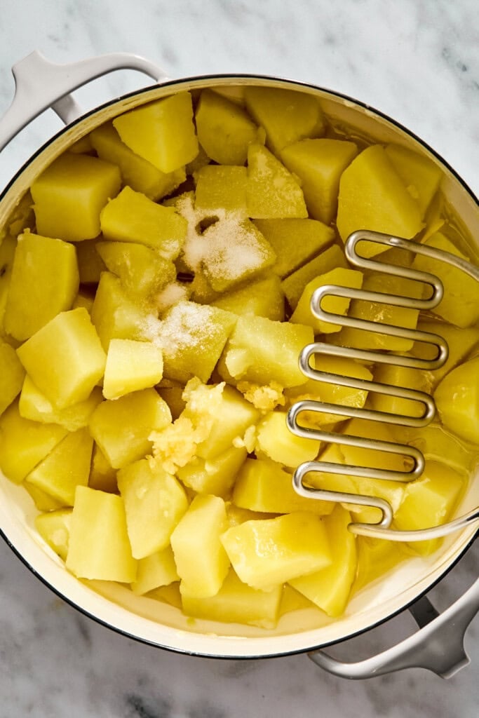 A metal potato masher resting in a pot of cubed boiled potatoes, sprinkled with salt and minced garlic.