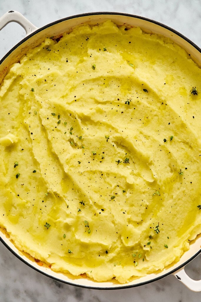 A stovetop pan filled with smooth mashed potatoes drizzled with oil and topped with herbs and black pepper.