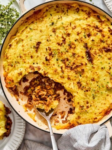 Shepherds pie in a shallow pan with a metal spoon scooping out a serving, topped with fresh herbs.
