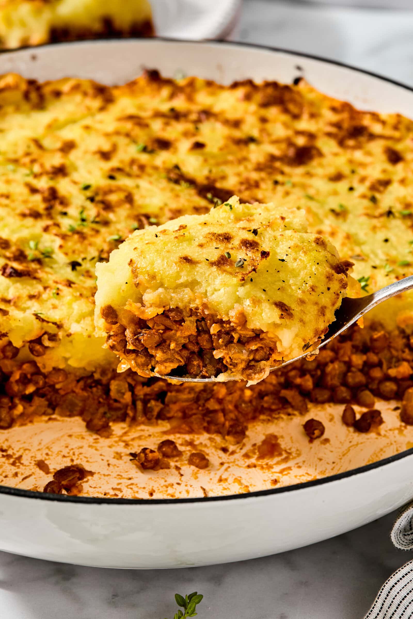 A spoonful of shepherds pie held above a white baking dish containing the rest of the meal, featuring a browned mashed potato crust over a layer of lentils.