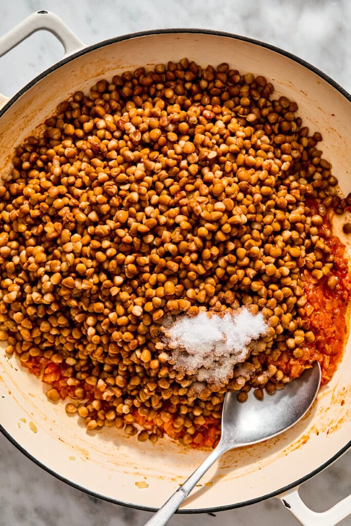 A large pan filled with cooked brown lentils, a mound of salt, and a spoonful of red sauce at the bottom.