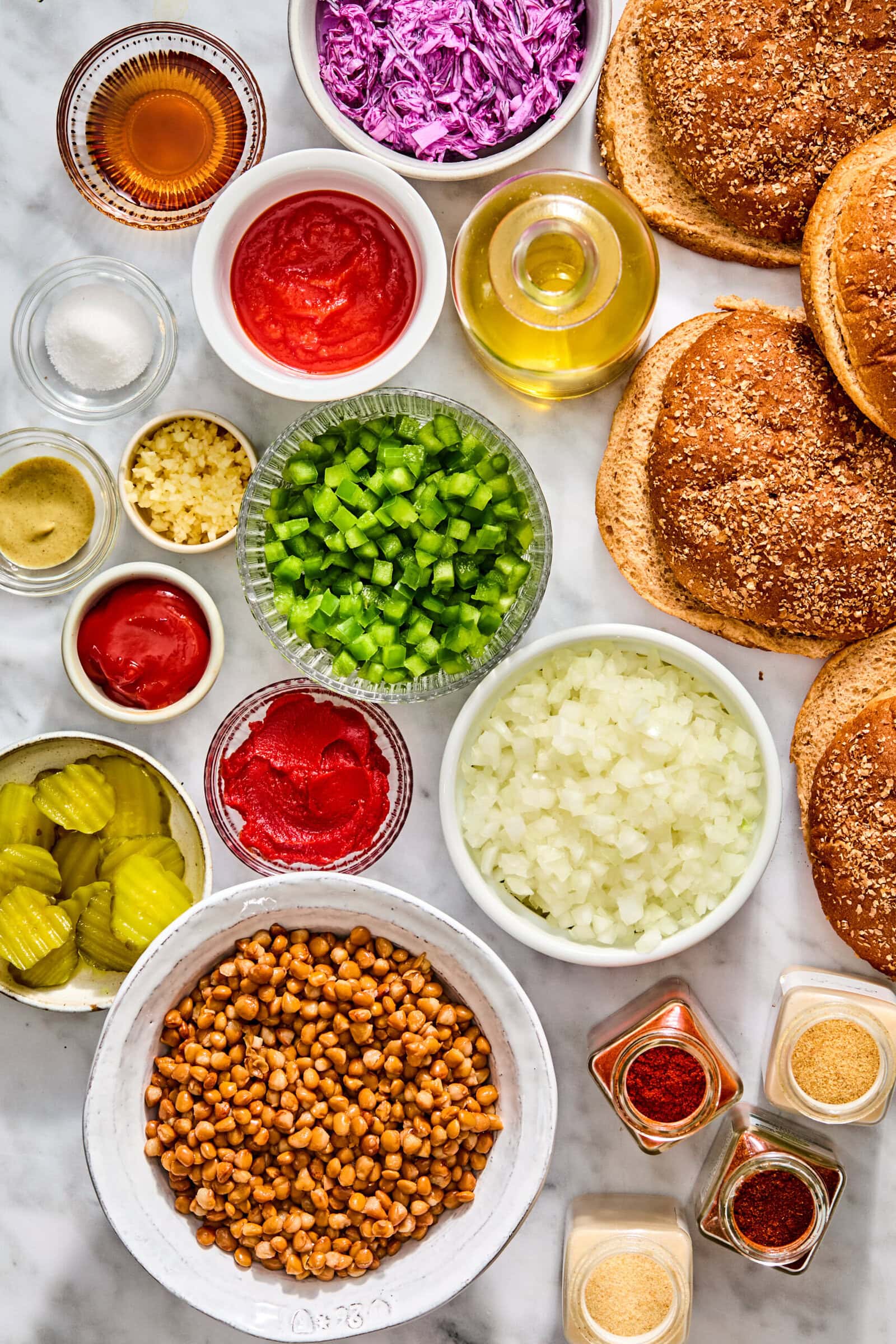 A top-down view of ingredients for cooking, including bowls of cooked lentils, chopped green peppers, onions, shredded cabbage, tomato paste, condiments, burger buns, and various spice jars.