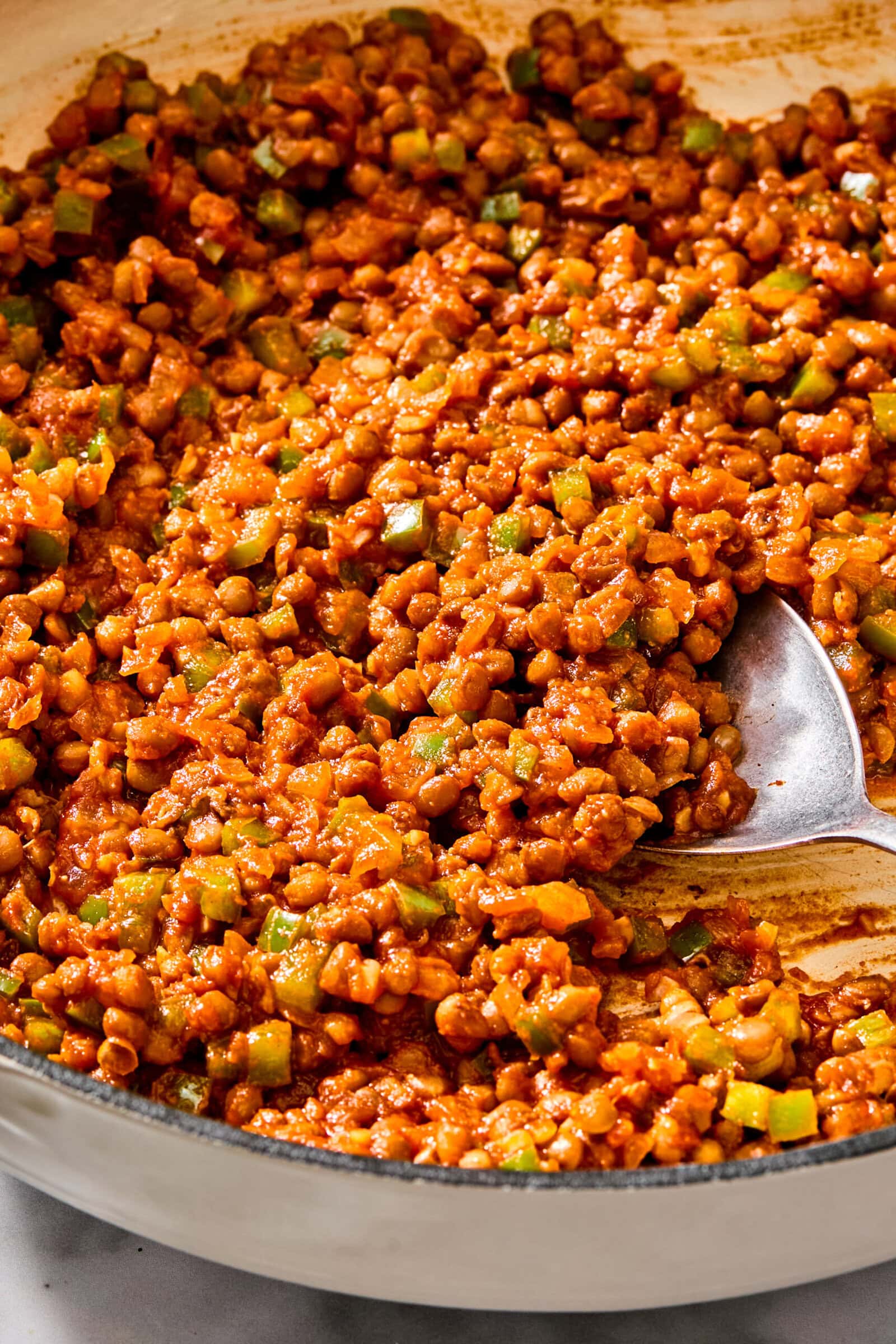 A skillet filled with a mixture of brown lentils, diced green peppers, and red tomato sauce, being stirred with a large metal spoon.