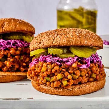 Two lentil sloppy joes on whole wheat buns, topped with shredded purple cabbage and pickle slices, on a wooden board.
