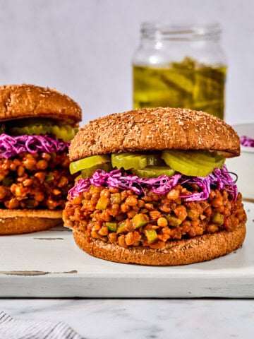 Two lentil sloppy joes on whole wheat buns, topped with shredded purple cabbage and pickle slices, on a wooden board.