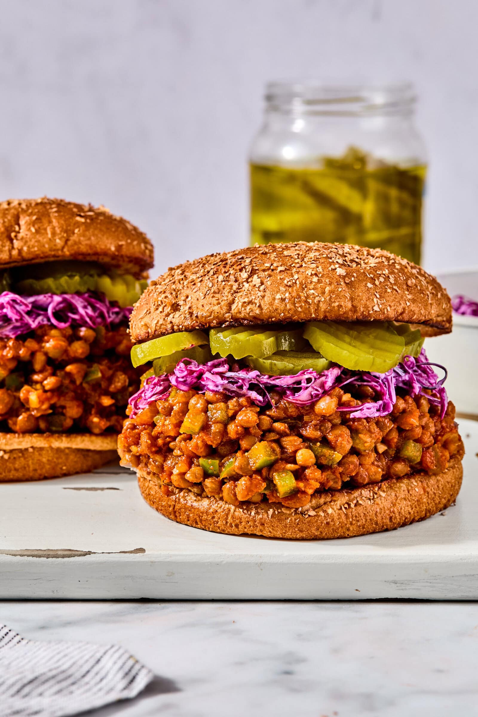 Two lentil sloppy joes on whole wheat buns, topped with shredded purple cabbage and pickle slices, on a wooden board.