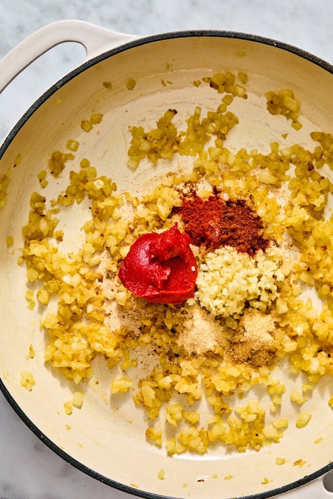 A skillet containing sautéed onions with piles of tomato paste, minced garlic, paprika, and ground spices.