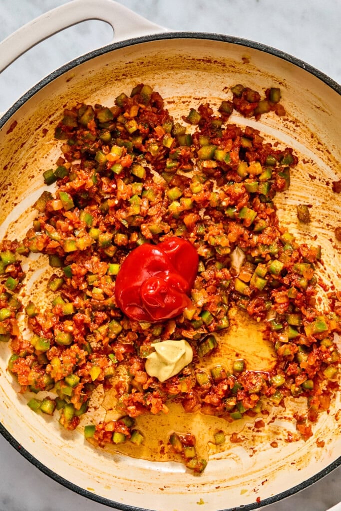 A dollop of ketchup and a dollop of mustard sit on top of a sautéed mixture of chopped vegetables in a wide skillet.