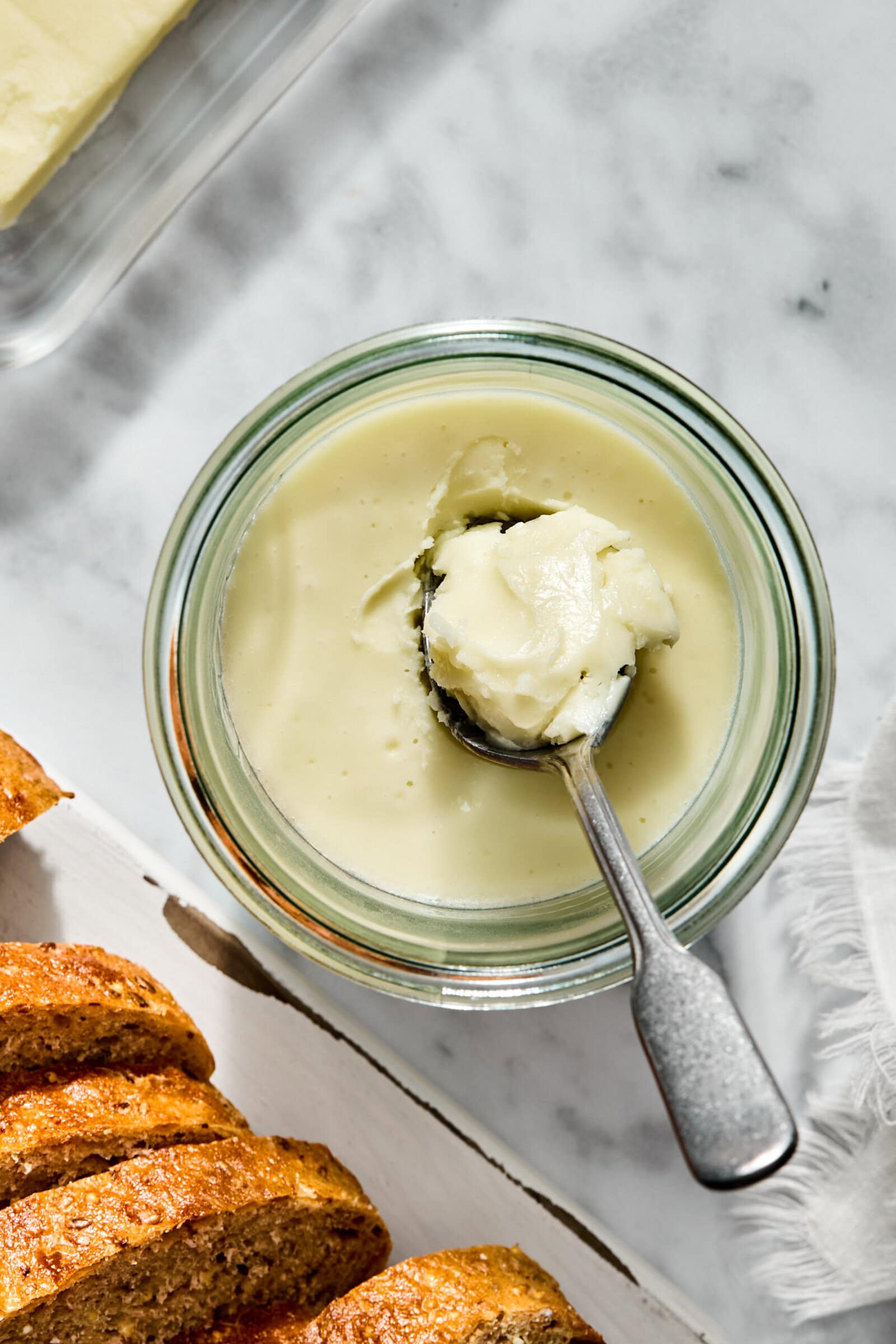 A spoonful of creamy butter in a glass jar, set beside slices of crusty bread.