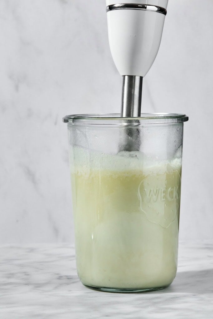 An immersion blender blending a light green liquid in a glass jar.