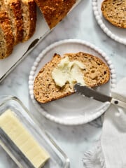 A slice of seeded bread on a small plate being spread with butter, next to a loaf and a glass butter dish.