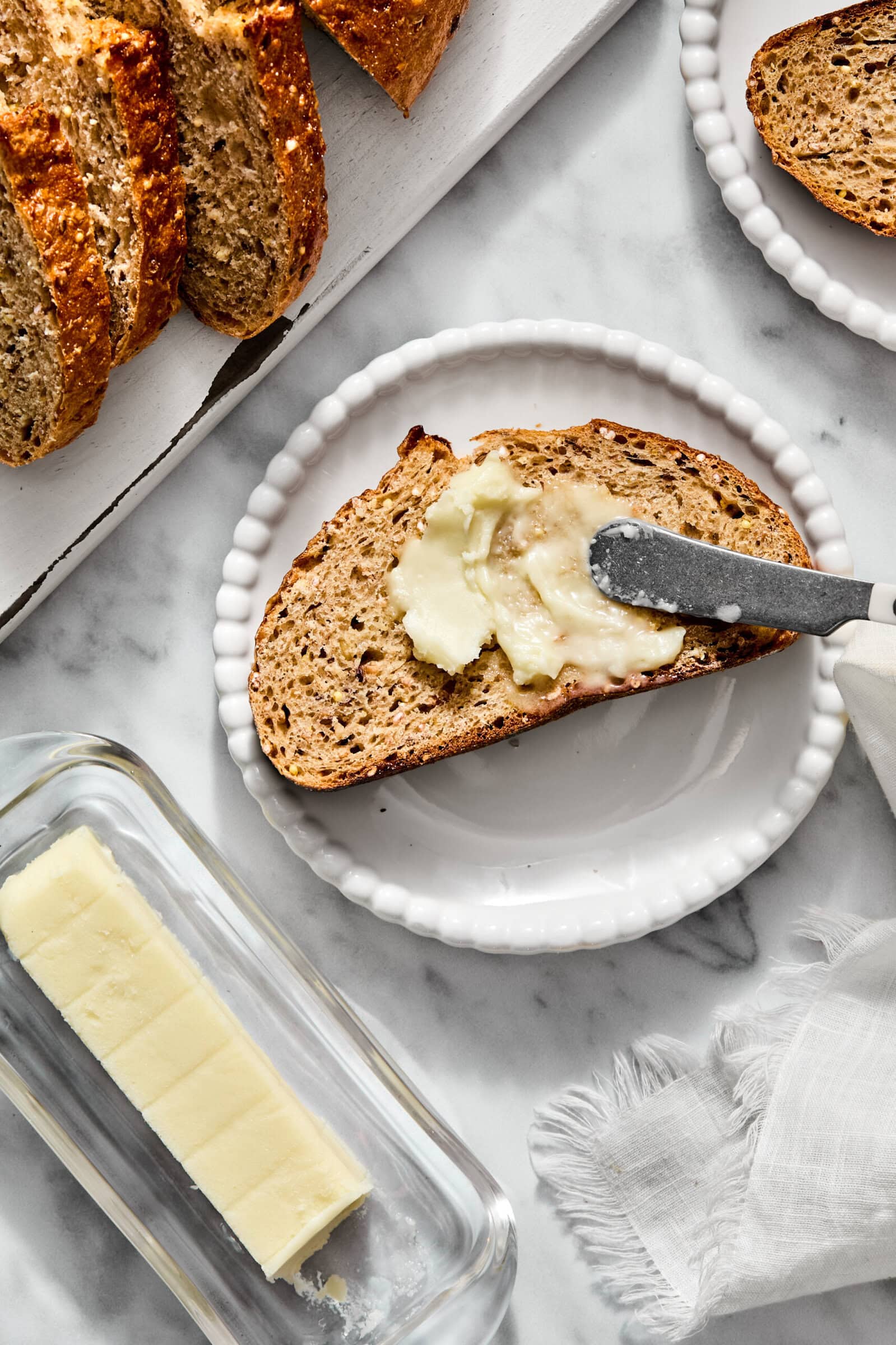 A knife spreads butter on a slice of whole grain bread on a white plate, with a loaf of bread and a stick of butter nearby.