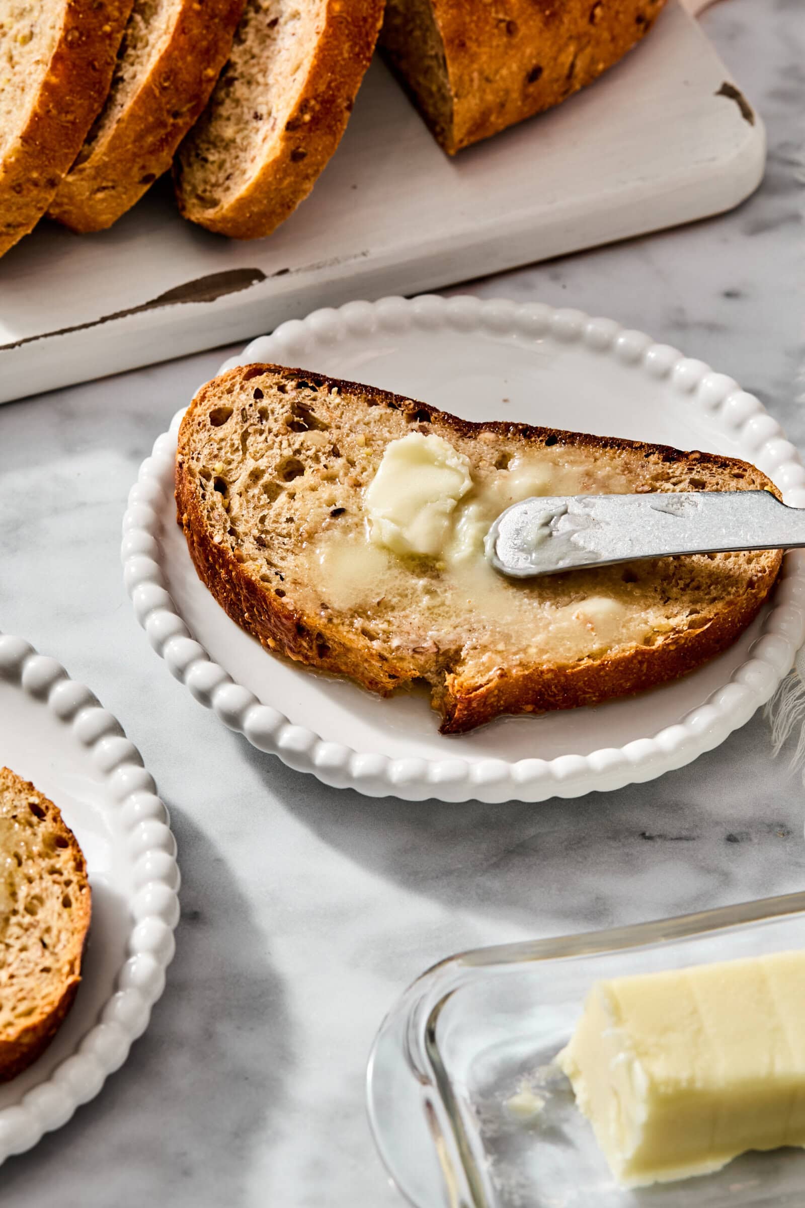A slice of toasted seeded bread on a small white plate being spread with butter using a silver knife, with additional bread and butter nearby.