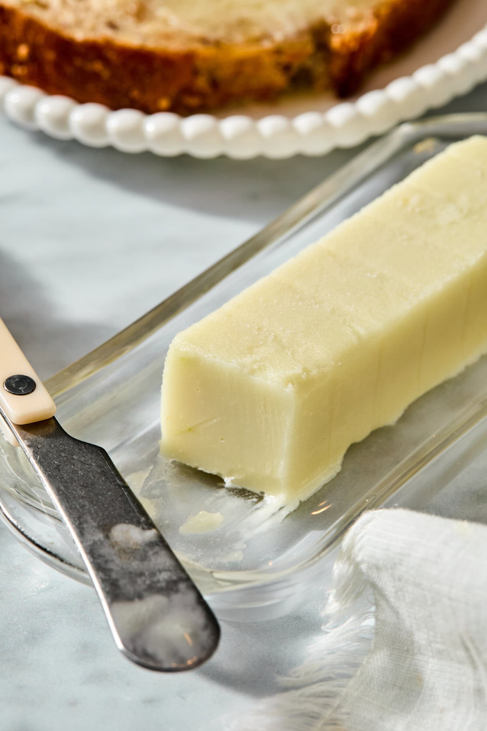 A stick of butter on a glass dish with a butter knife and a slice of toasted bread in the background.