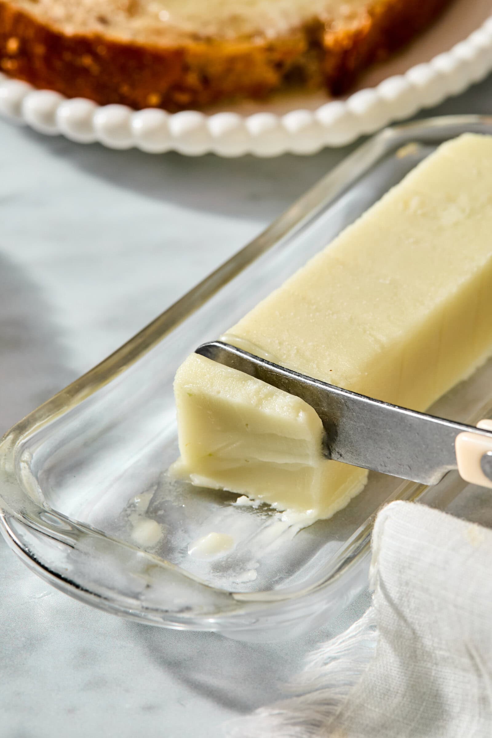 A knife cutting a slice from a stick of butter in a glass dish.