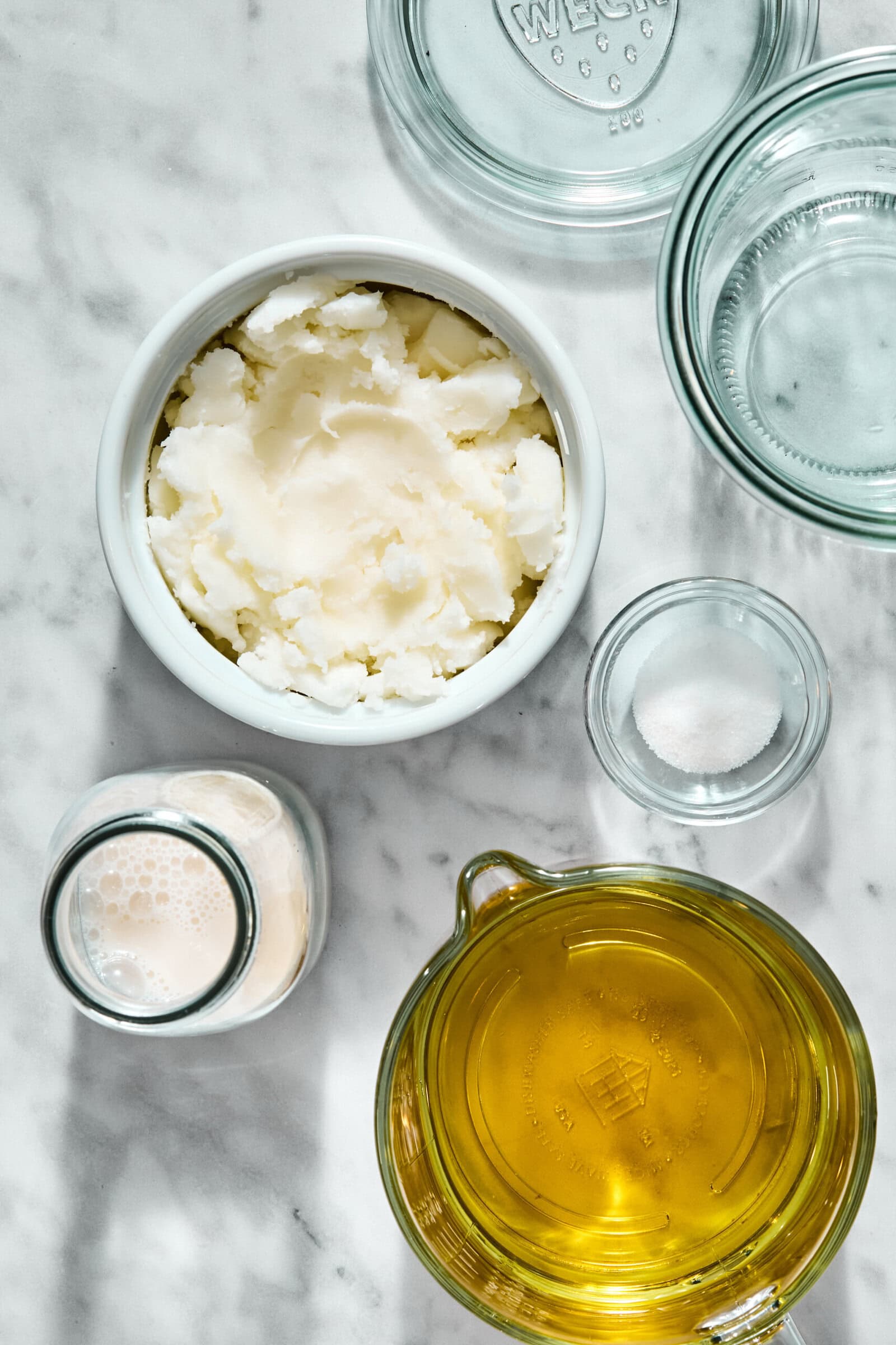 A top-down view of ingredients on a marble surface, including a bowl of solid fat, a pitcher of oil, a small glass of milk, a small dish of salt, and glass lids.