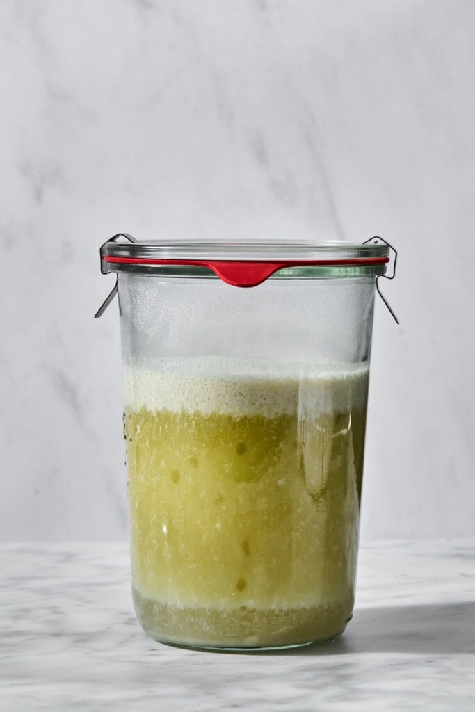 A sealed glass jar containing a mixture that has separated into a cloudy green liquid at the bottom and a layer of white foam at the top.