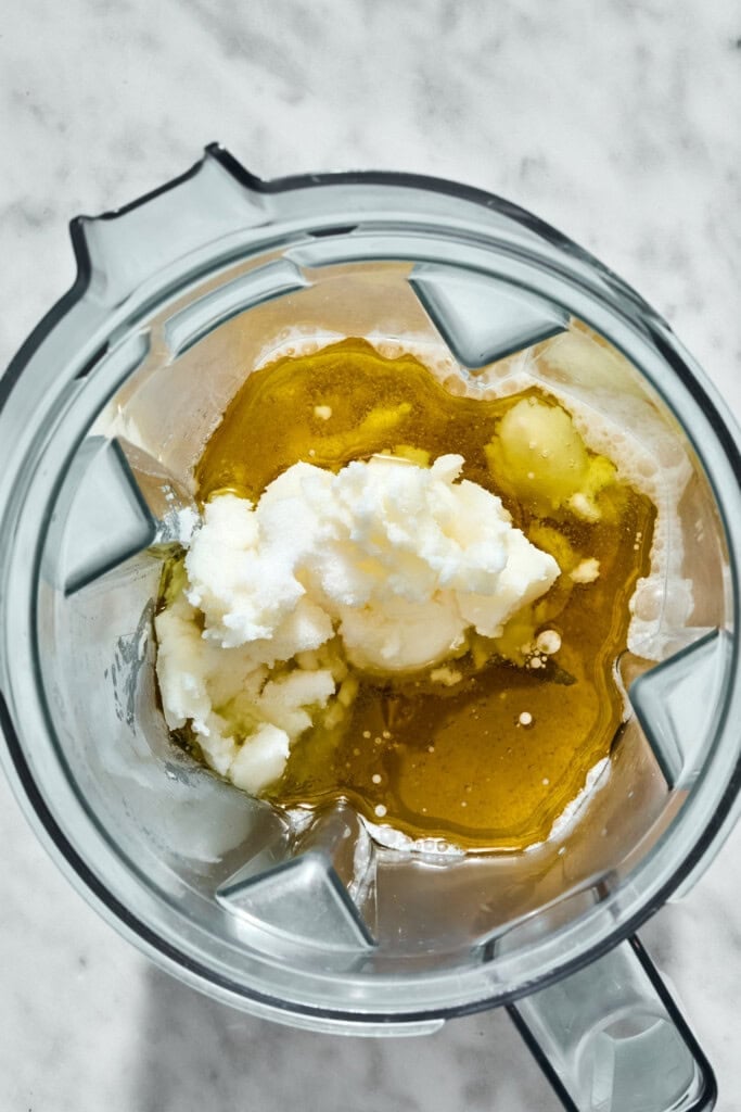 A blender jar containing a mixture of golden oil and a scoop of white, granular solid.