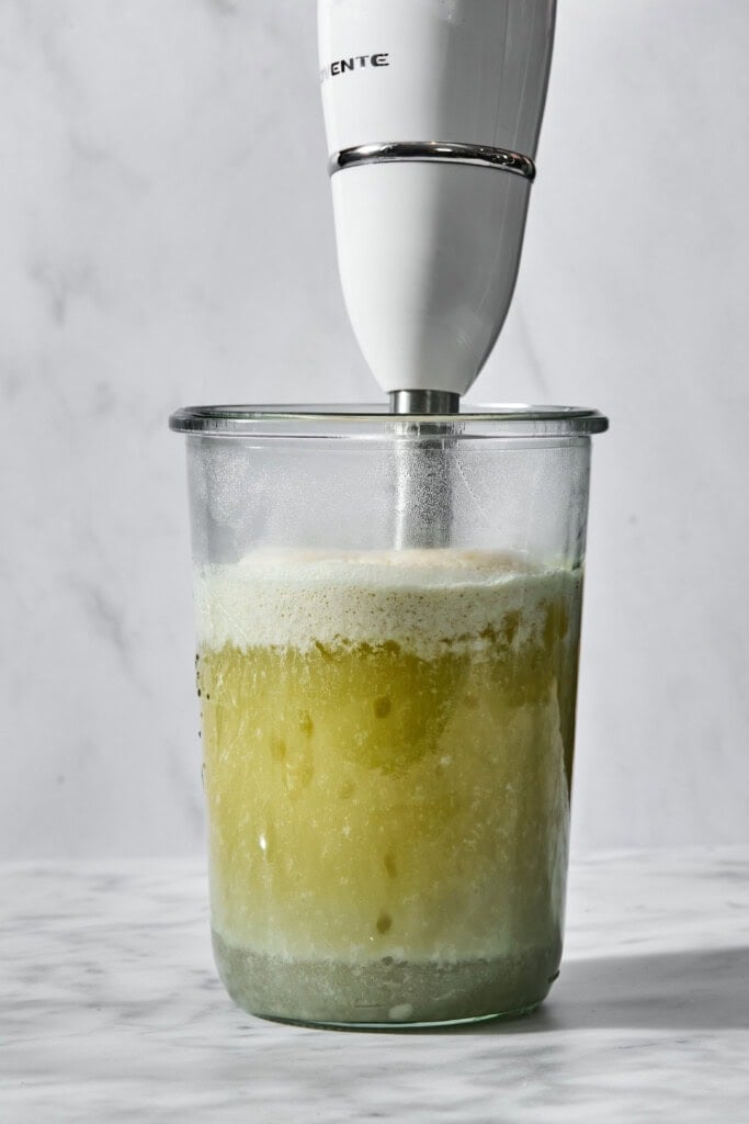 An immersion blender in a clear glass container blending a pale green, foamy liquid.